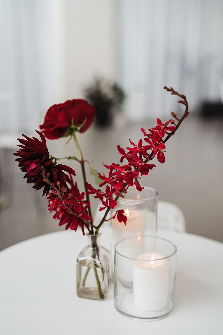 two vases with red flowers and candles on a white table in front of a window