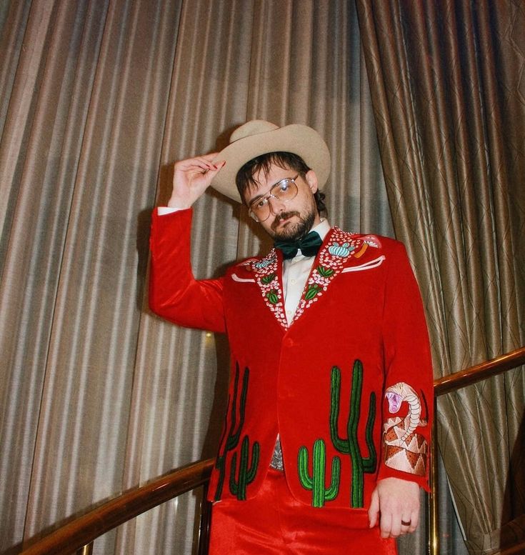 a man in a red suit and hat standing next to a stair case with curtains behind him