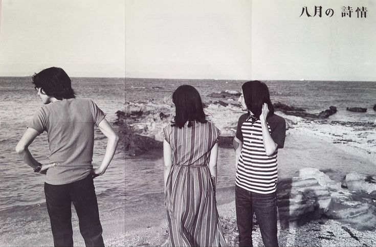 three people standing on the beach looking out at the water and rocks in front of them