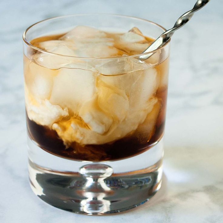 a glass filled with ice and liquid on top of a marble counter next to a metal spoon