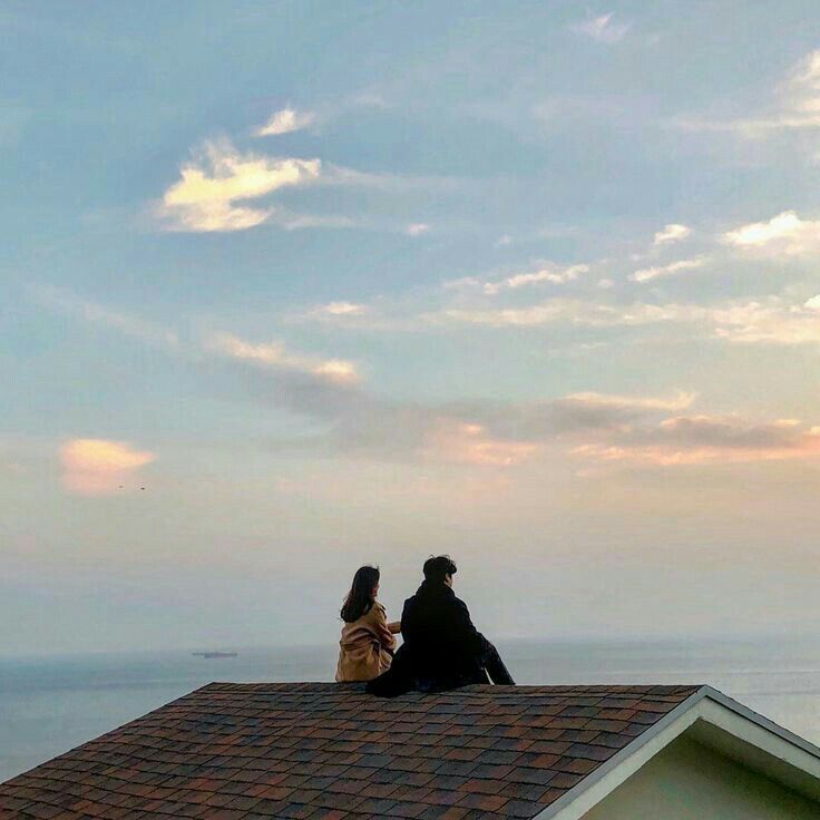 two people sitting on the roof of a house looking out at the ocean and sunset