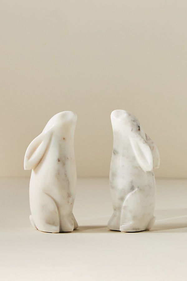 two marble rabbits sitting next to each other on a white surface with a light colored wall in the background