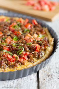 a quiche with meat and vegetables in a pie pan on a wooden table next to a cutting board