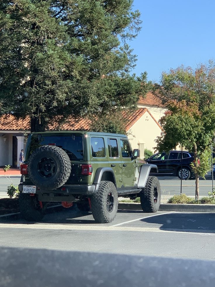 a green jeep parked in a parking lot next to a tree and building with cars behind it