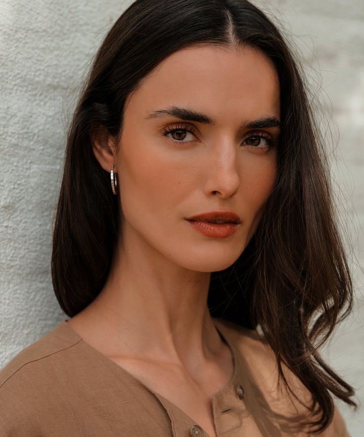 a woman with dark hair wearing earrings and a brown shirt is standing against a white wall