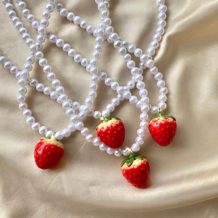 three necklaces with strawberries and pearls on a white cloth background, one is made out of plastic beads