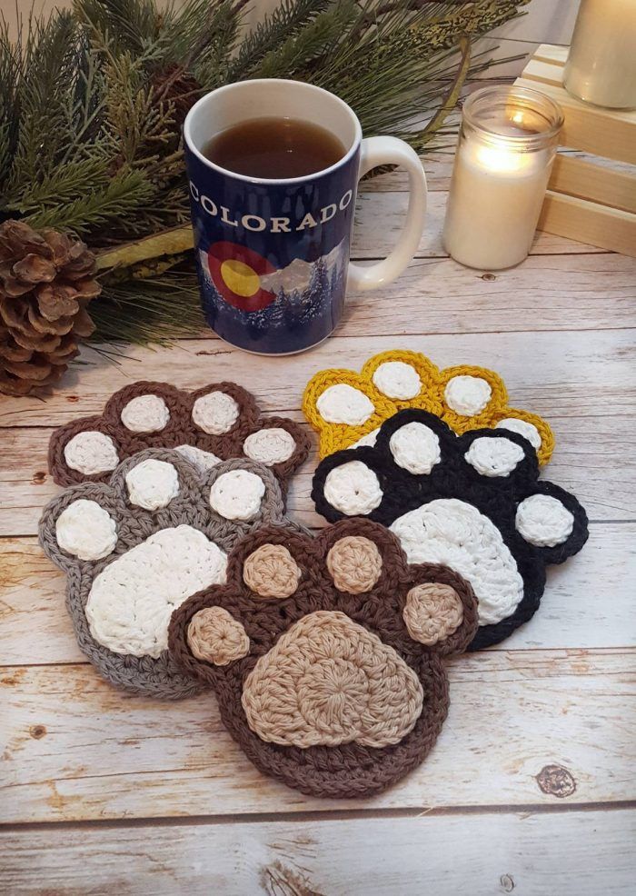 four crocheted coasters with paw prints next to a cup of tea and pine cones