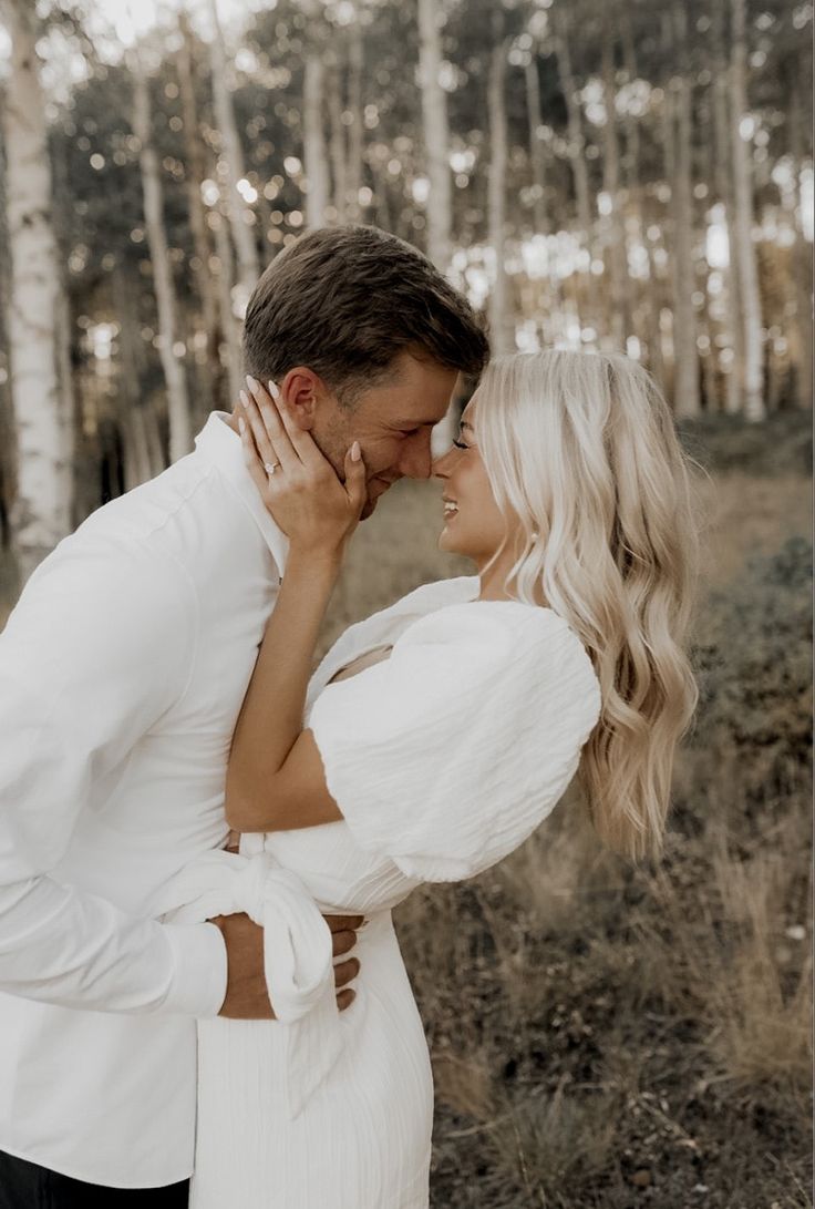 a man and woman standing next to each other in front of some trees with their faces close together