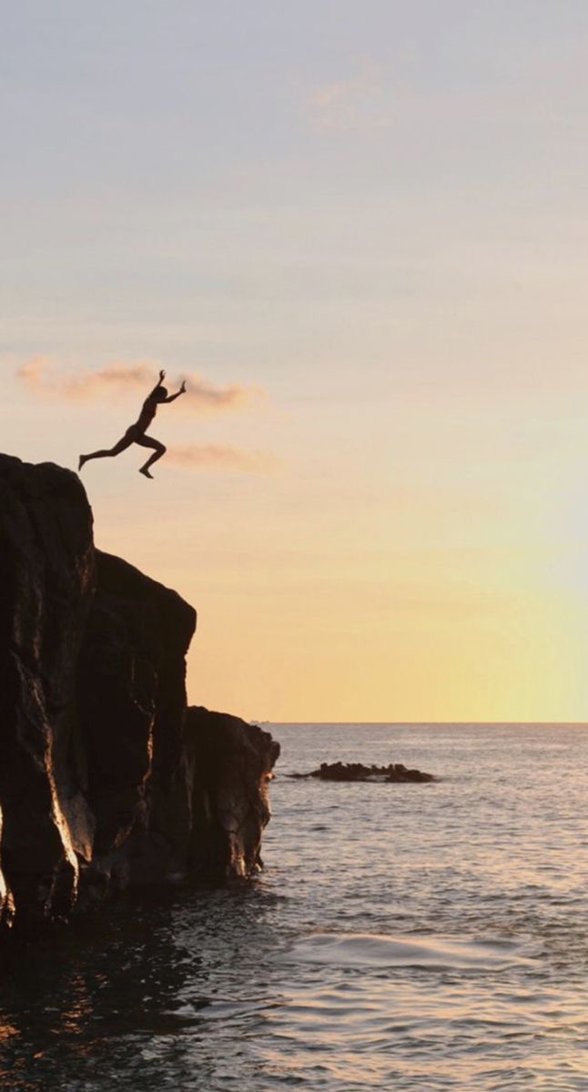 a person jumping off the edge of a cliff into the ocean at sunset or dawn