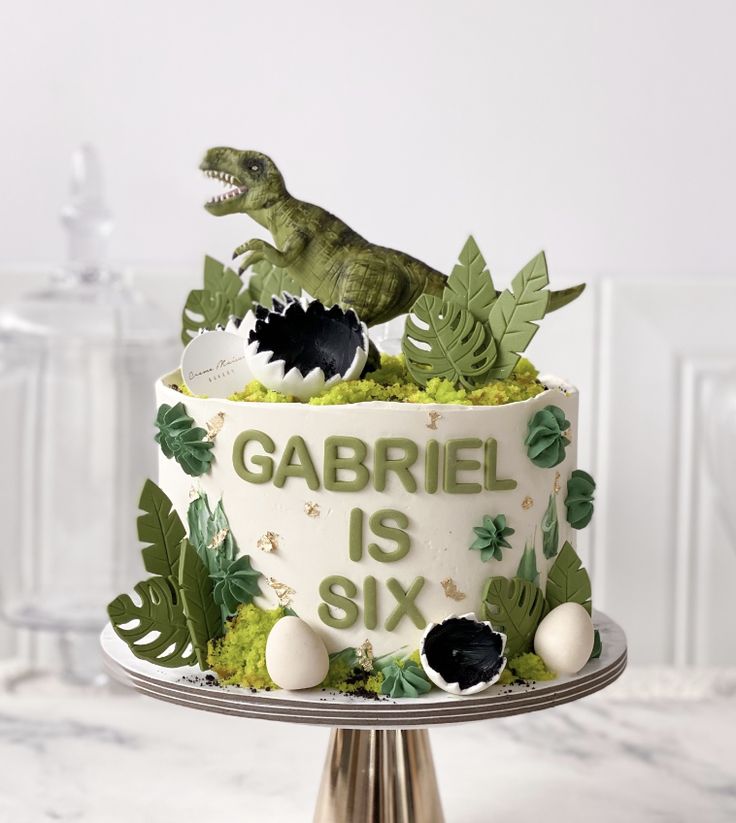 there is a dinosaur cake on top of a stand with plants and balls around it