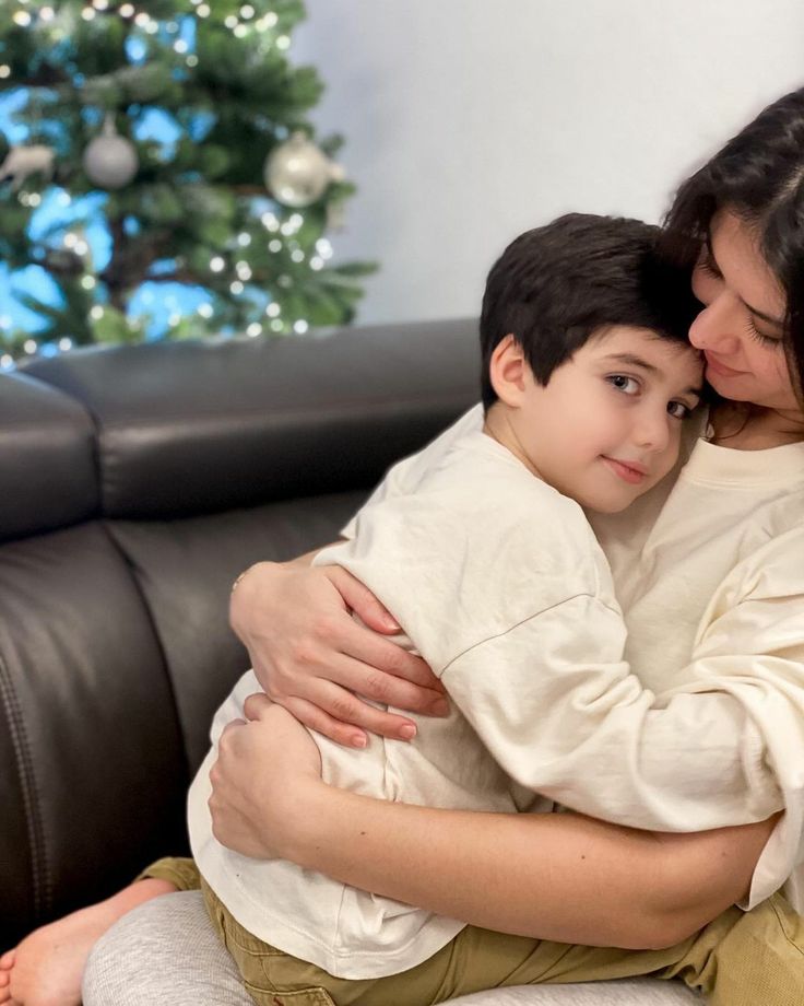 a woman hugging a boy on the couch with a christmas tree in the back ground