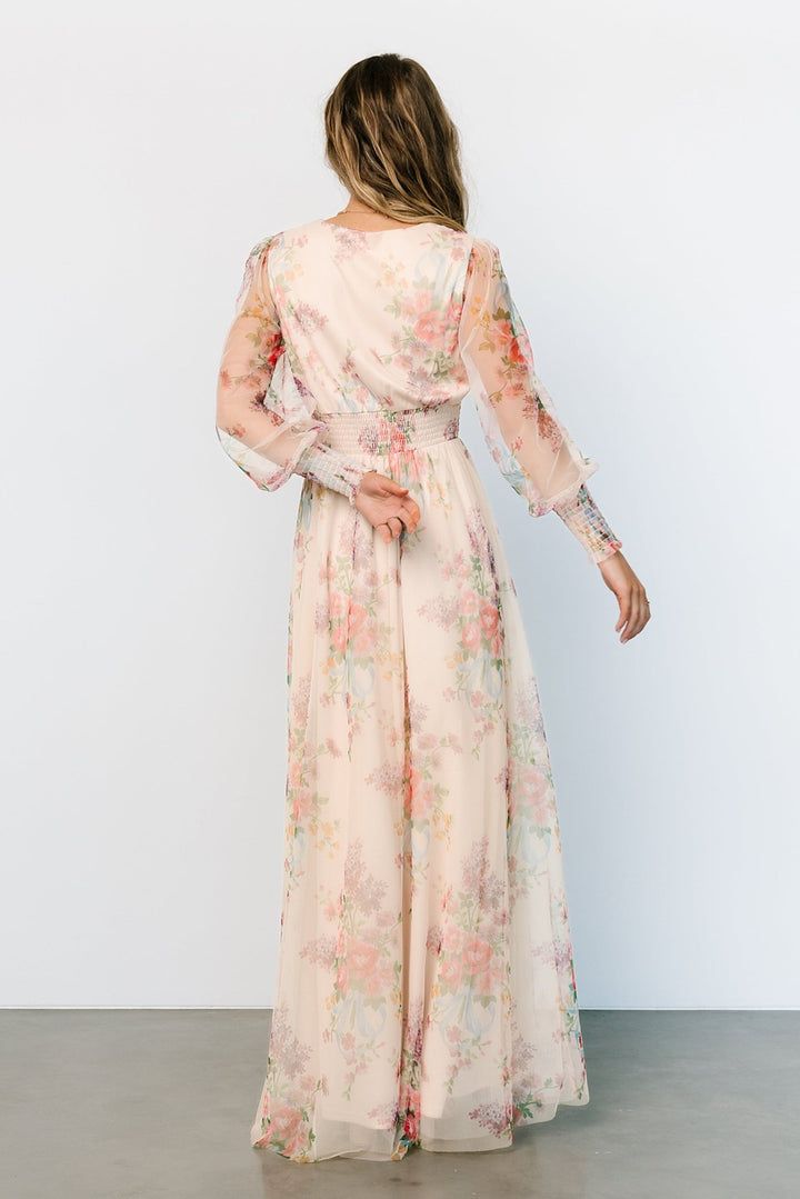 a woman wearing a long dress with floral print on the sleeves and back, standing in front of a white wall