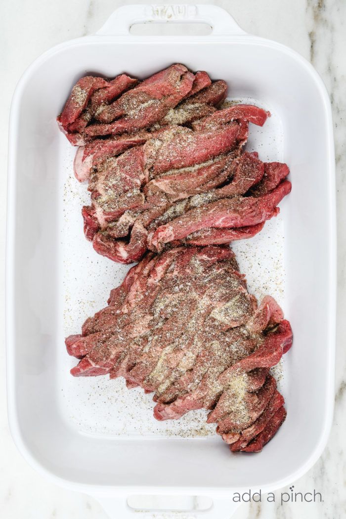 raw steaks in a white casserole dish on a marble counter top with seasoning sprinkles