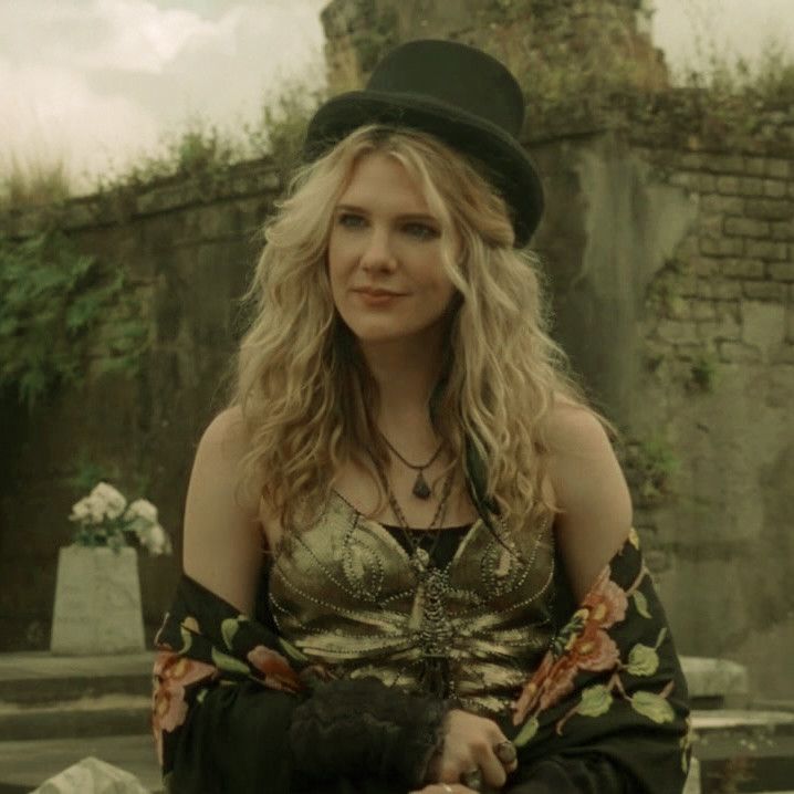 a woman with long blonde hair wearing a black hat and dress sitting on the ground