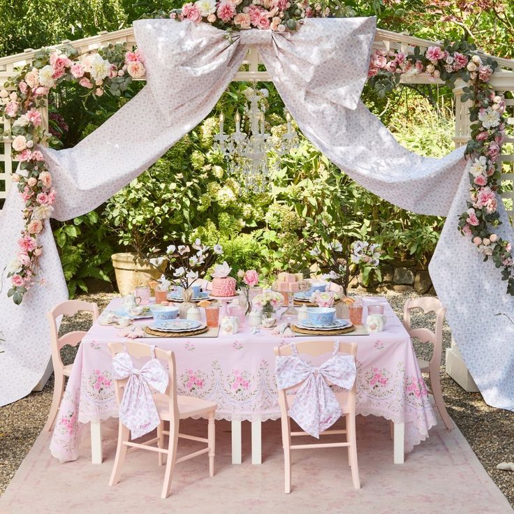 a table set up for a tea party with pink flowers on the top and white drapes