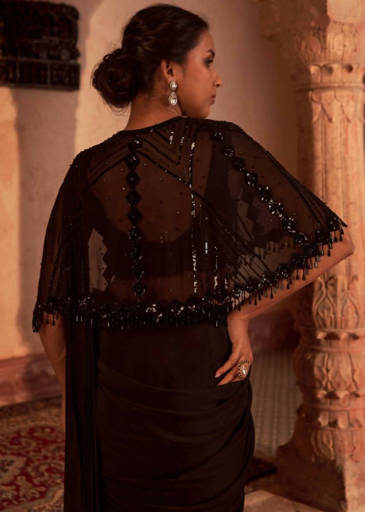 Elegant drape sari in crepe is paired with fully embroidered sequins and beaded georgette cape. The outfit is paired with padded silk blouse and embroidered belt. Designer Pre-draped Saree With Cape Sleeves For Eid, Festive Black Sets With Cape Sleeves, Festive Black Set With Cape Sleeves, Festive Black Cape Sleeve Sets, Glamorous Georgette Dresses For Diwali, Black Sets With Cape Sleeves For Evening, Black Evening Sets With Cape Sleeves, Black Evening Set With Cape Sleeves, Georgette Cape Sets