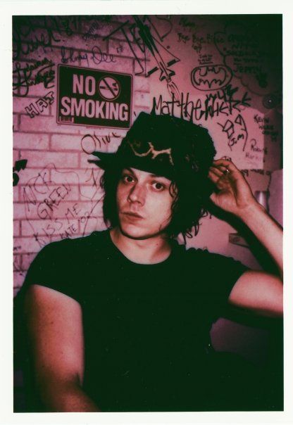 a man wearing a hat and glasses in front of a brick wall with graffiti on it