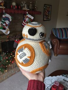 a hand holding a knitted bbg ball in front of a christmas tree