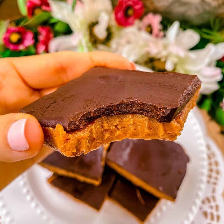 a person holding up a piece of chocolate to show it's side on a plate with flowers in the background