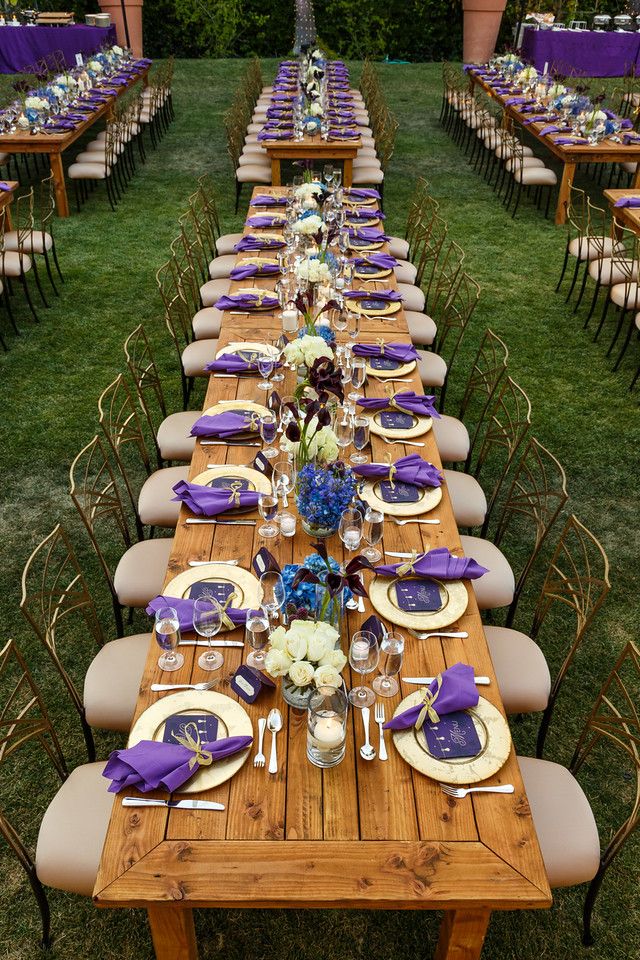 a long wooden table with purple and white napkins on it is set for an outdoor dinner