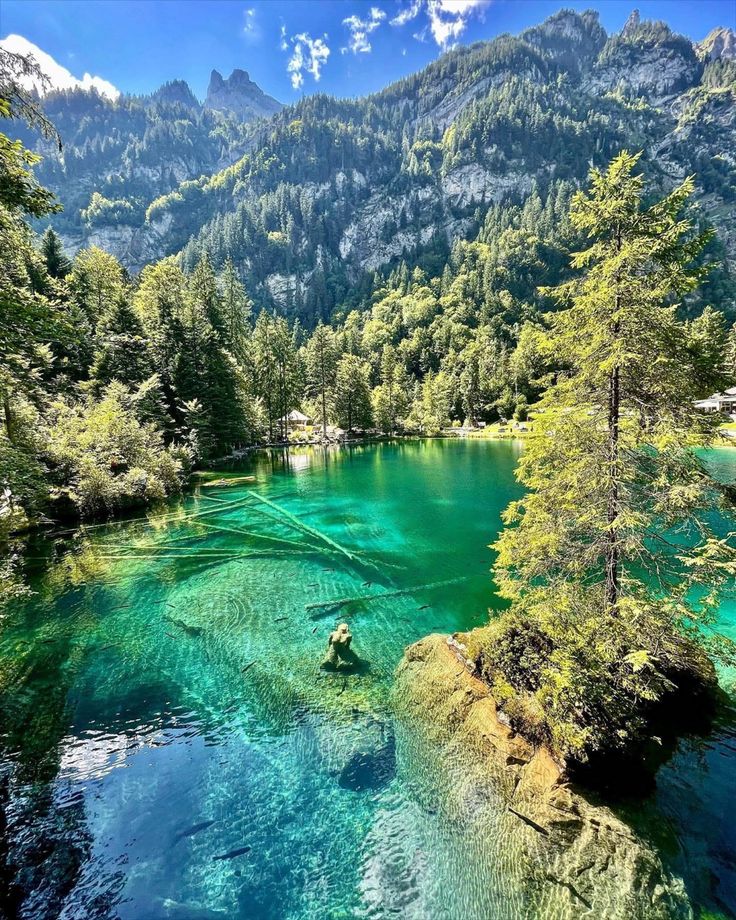 a lake surrounded by mountains and trees with blue water in the foreground, on a sunny day