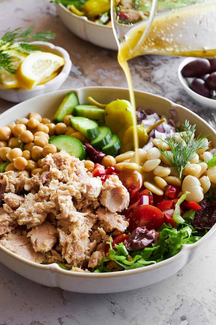 someone pouring dressing onto a salad in a white bowl on a marble countertop next to other dishes