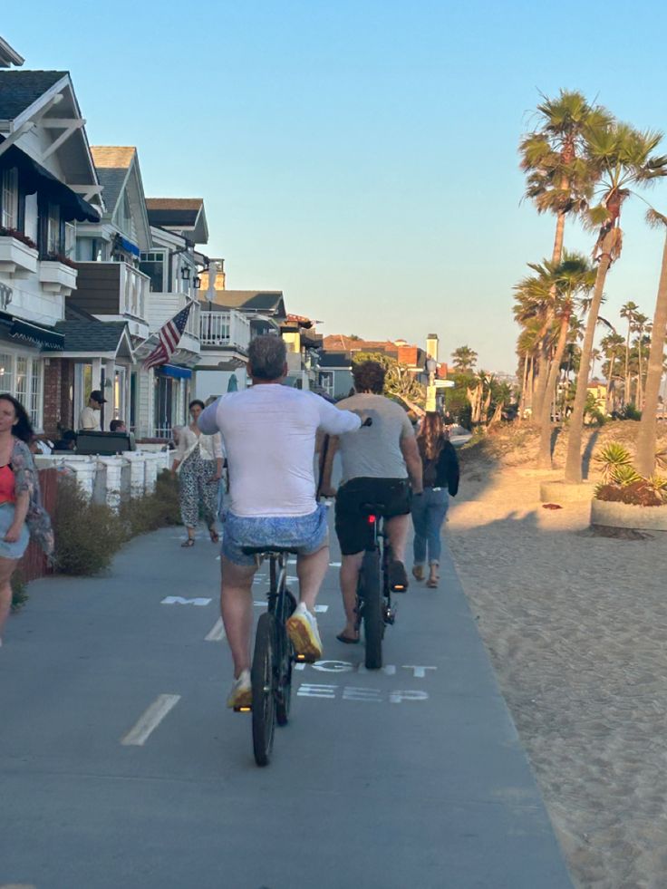 people are riding bikes on the sidewalk near beach houses with palm trees in the background