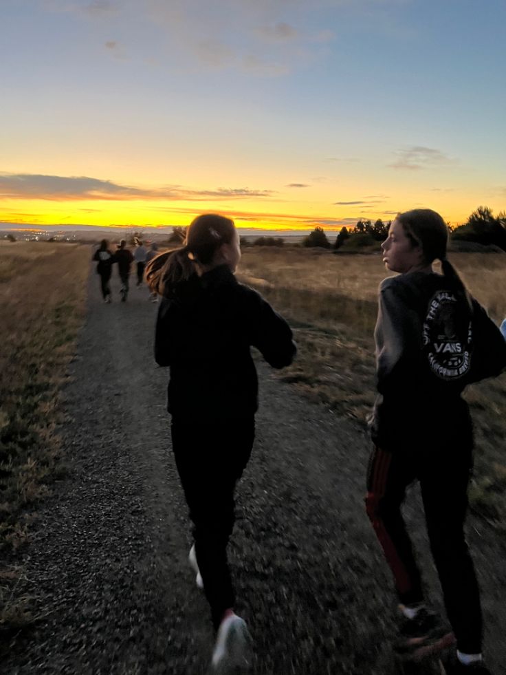 two girls running down a dirt road at sunset with the sun setting in the background