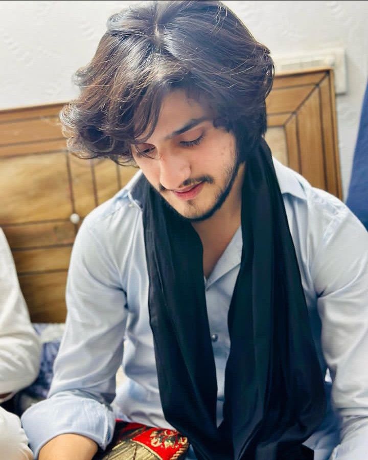 a man with long hair wearing a scarf and looking at his cell phone while sitting on a bed