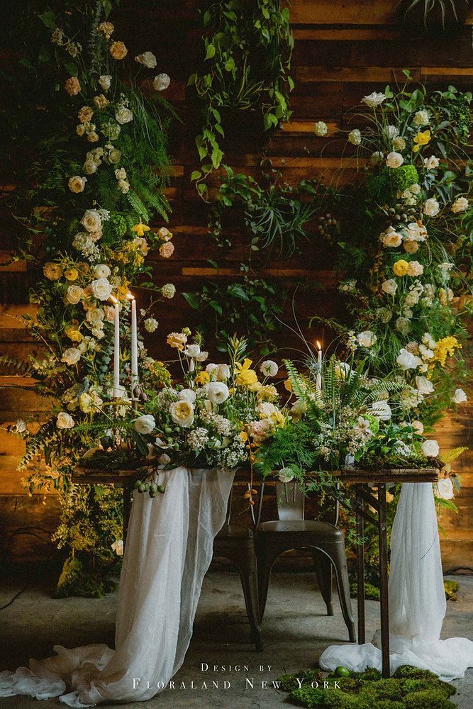 a table with flowers and candles on it in front of a wooden wall filled with greenery