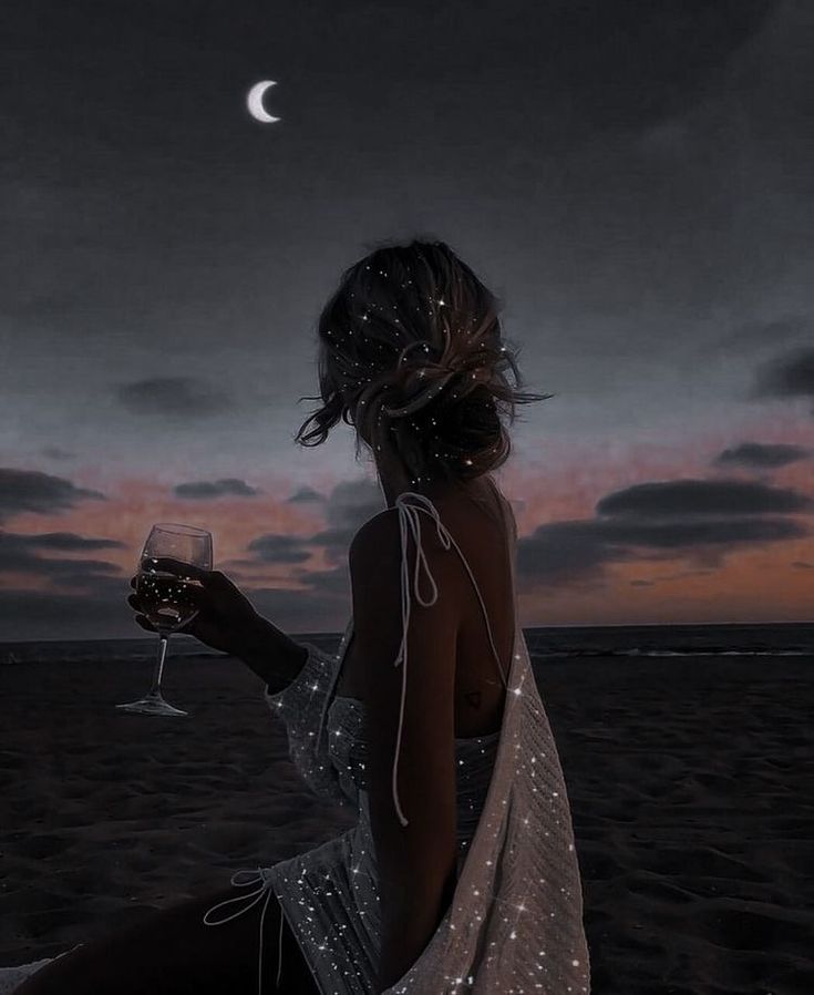 a woman sitting on the beach with a glass of wine in her hand at night