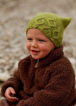 a young boy wearing a green knitted hat and coat sitting on the ground with his hands in his pockets