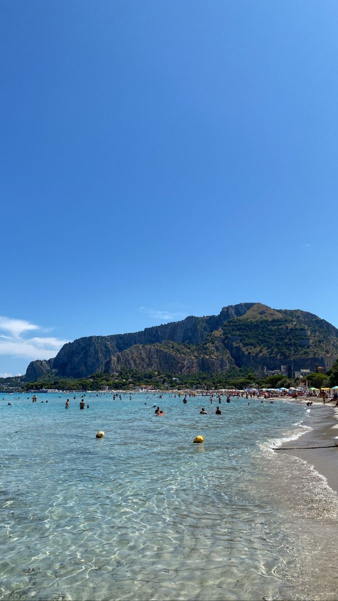 many people are swimming in the clear blue water on a beach with mountains in the background