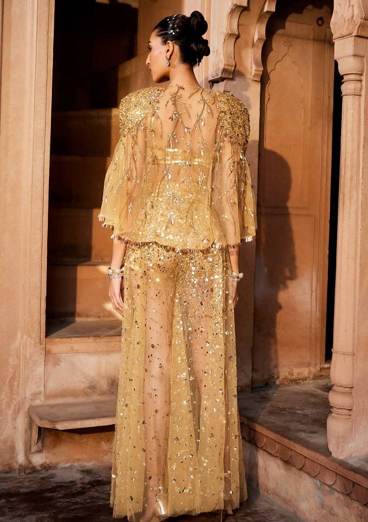 A bold three piece dress in sheer fabric, a glamorous embellished gold bralet paired with sheer trousers with splatter embroidery of sitara/patra and a matching sheer jacket with power shoulder highlighted with gold embellishments Glamorous Gold Hand-embellished Sharara, Glamorous Gold Hand Embellished Sharara, Hand Embellished Sharara For Eid Party, Glamorous Gold Hand-embellished Sets, Eid Party Hand Embellished Sharara, Hand Embellished Evening Sets For Eid, Elegant Evening Sets For Festivals, Hand Embellished Sharara For Evening Party Wear, Glamorous Gold Embellished Sets