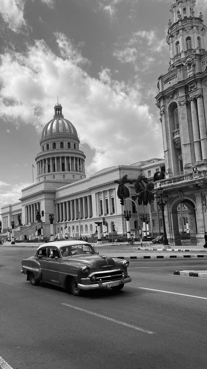 an old red car is driving down the street in front of a large white building