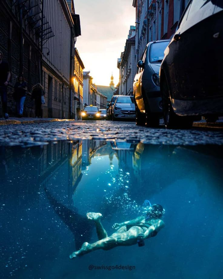 a man swims in the water next to parked cars on a narrow city street