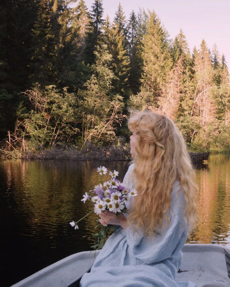 a woman with long hair sitting in a boat on a lake holding flowers and looking at the water