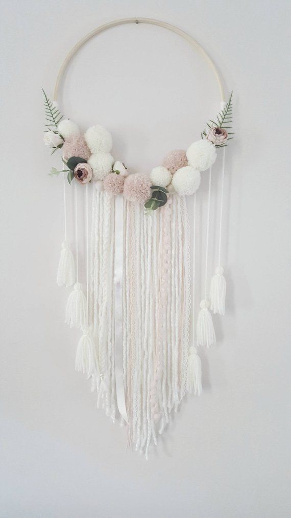 a white circular wall hanging with flowers and tassels on the sides, along with greenery