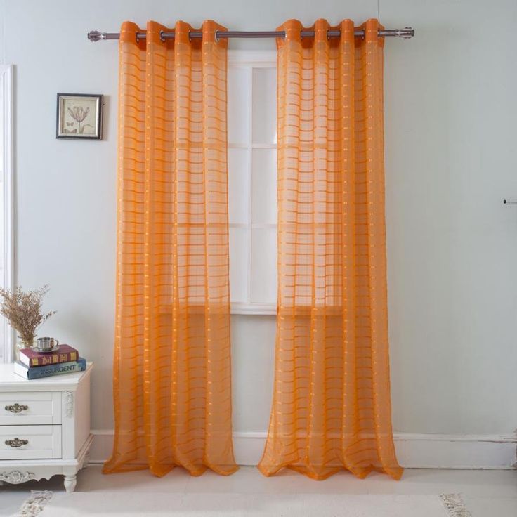 an orange sheer curtain hangs in front of a white dresser and window with pictures on the wall