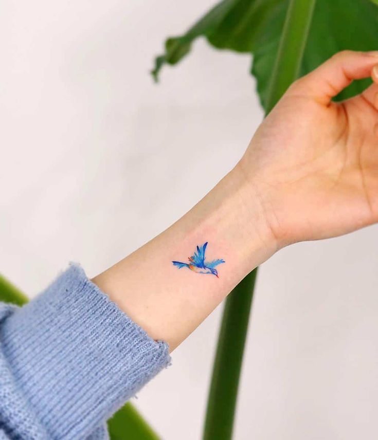 a small blue bird tattoo on the left inner arm and wrist, sitting next to a large green plant