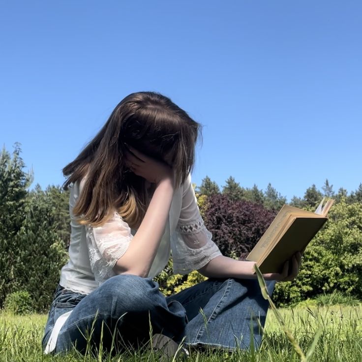 a woman sitting in the grass reading a book with her hands on her head and eyes closed