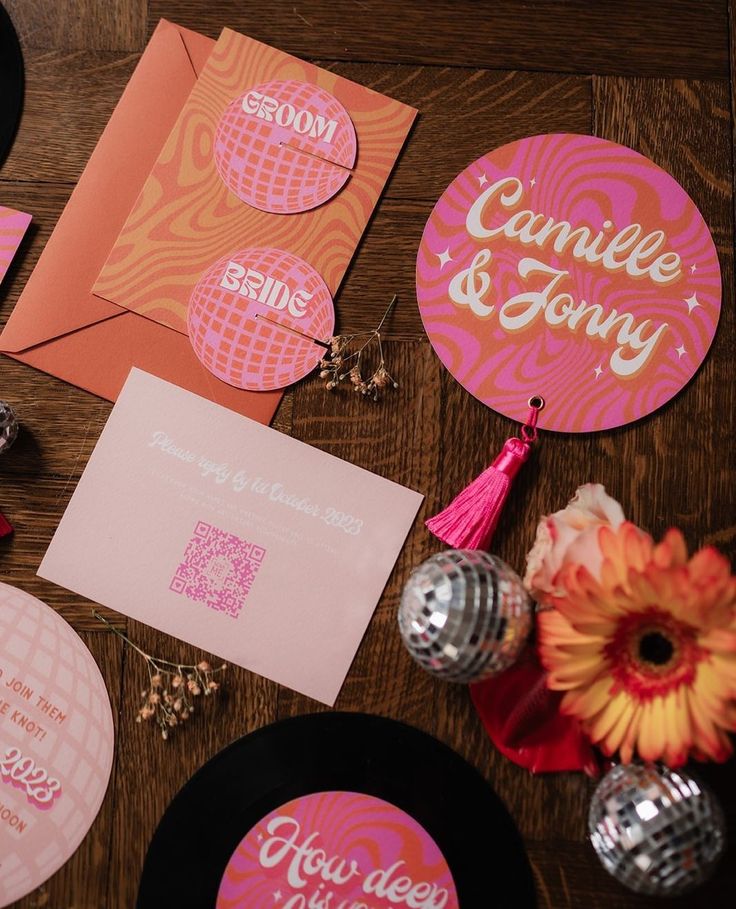 pink and orange wedding stationery on a wooden table with flowers, cards, and disco balls