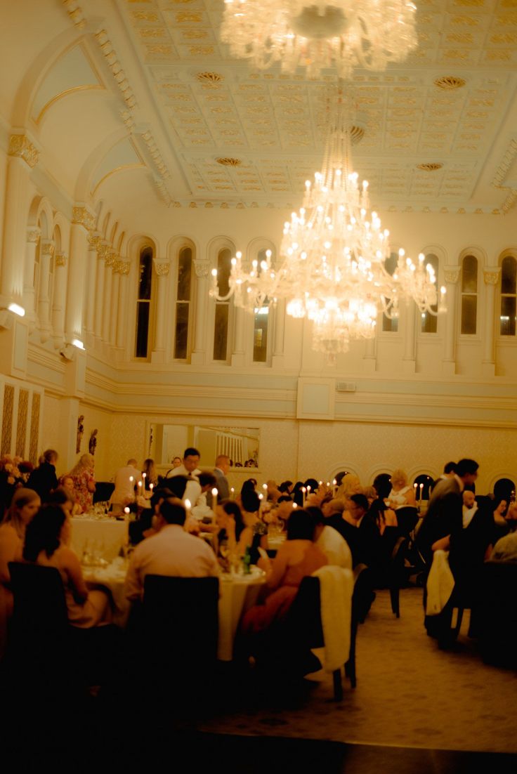 people are sitting at tables in a large room with chandeliers hanging from the ceiling