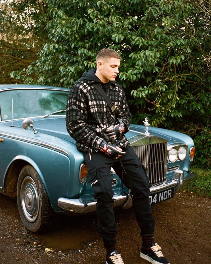 a man sitting on the hood of a blue car in front of some trees and bushes