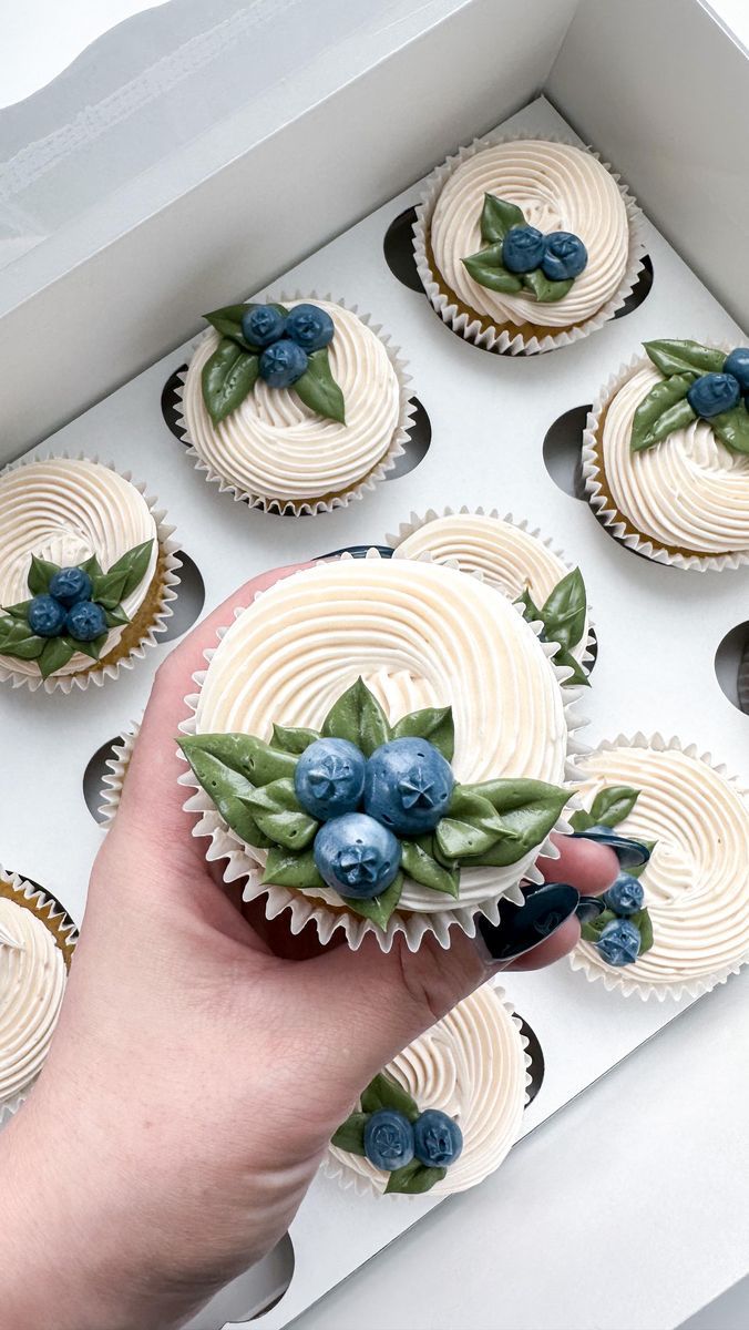 a hand is holding some cupcakes with blueberries and leaves on them in a box