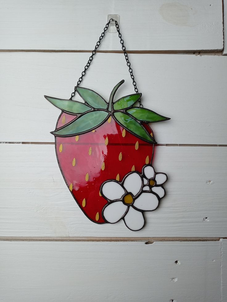 a red strawberry hanging from a chain on a white wooden wall with flowers in the foreground