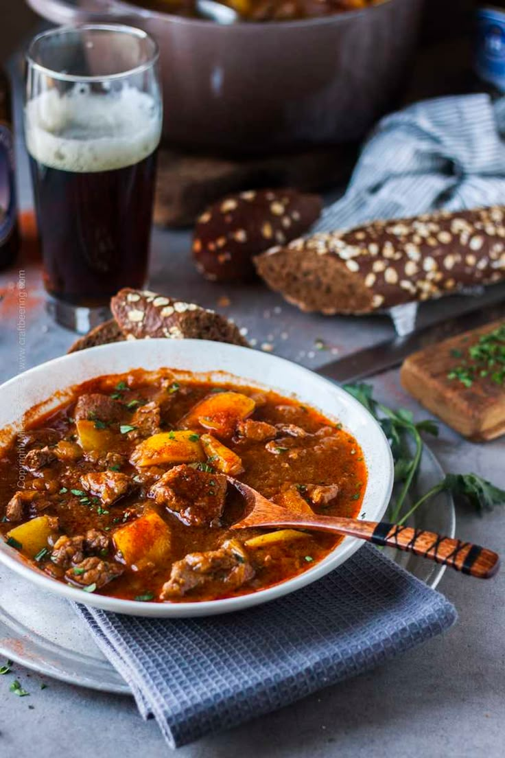 a bowl of stew with bread and beer in the background