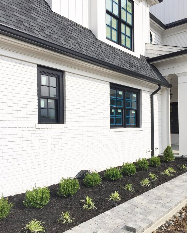 a white brick house with black trim and green plants on the front lawn, next to a walkway