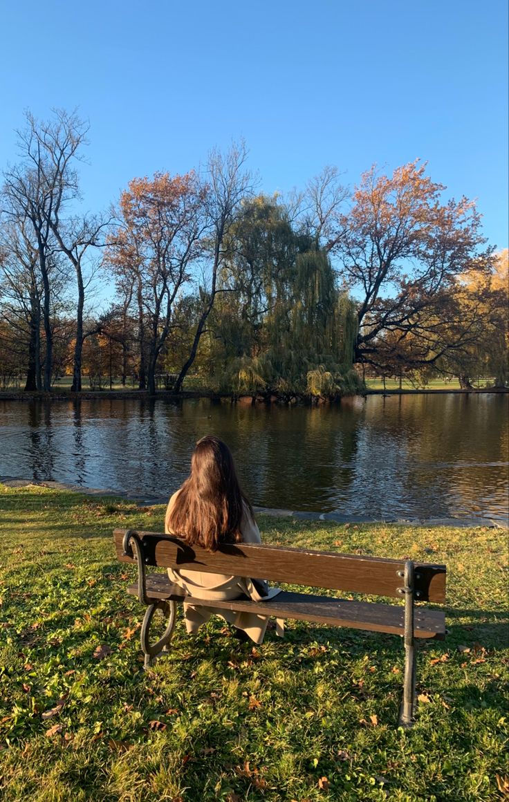 a woman is sitting on a bench in front of a lake and trees with no leaves