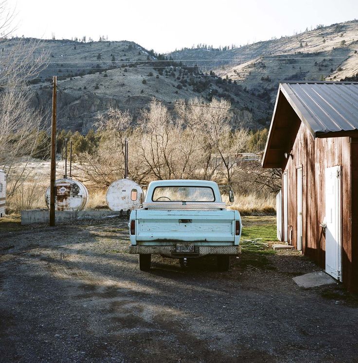 an old blue truck parked in front of a barn and some buildings with mountains in the background